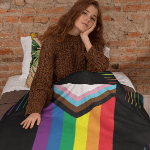 girl in brown jumper in bed with a blanket with the pride flag. Brick wall behind her, leaning on colorful pillows